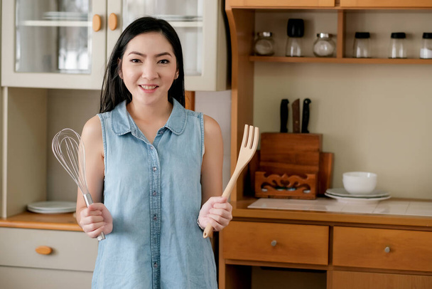 Asian woman hold whisk and fork stand in kitchen with day light and she look happy. Concept of preparation for cooking in her home. - Fotó, kép