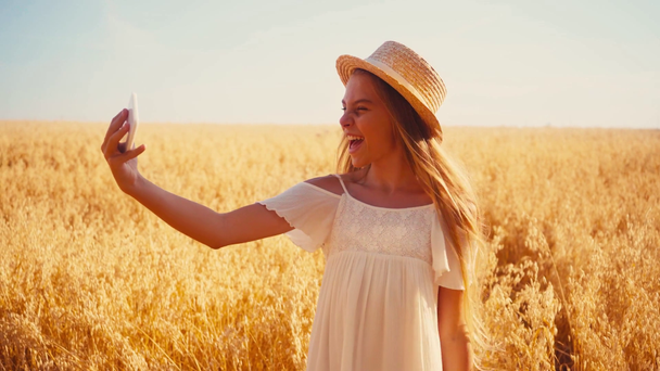 fröhliches Mädchen in weißem Kleid und Strohhut macht Selfie im Weizenfeld  - Filmmaterial, Video