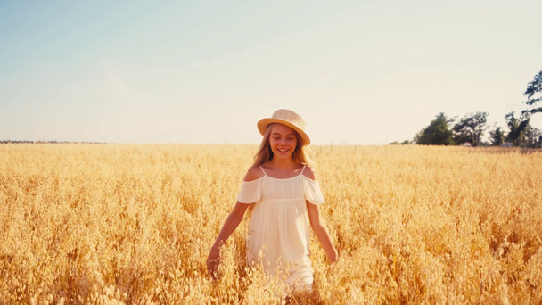 vrolijk meisje in witte jurk en stro hoed wandelen in het veld en aanraken strohoed - Video