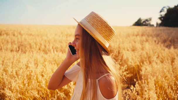 παιδί με λευκό φόρεμα και ψάθινο καπέλο μιλάει στο smartphone στον τομέα του σιταριού - Πλάνα, βίντεο