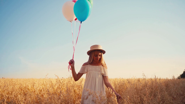 Beyaz elbiseli ve hasır şapkalı çocuk buğday tarlasında balonlarla yürüyor. - Video, Çekim