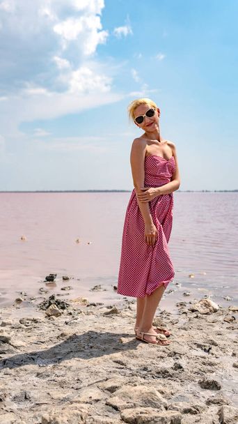 Joyful adult in red dress looking at camera on background of pink lake blue clouds during day and summer - Foto, Bild