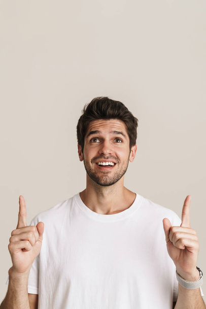 Image of joyful young man smiling and pointing fingers upward isolated by beige background - Fotoğraf, Görsel