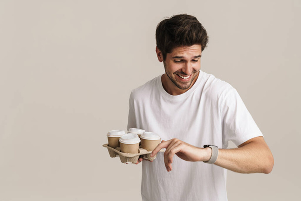 Image of joyful young man holding paper cups and looking at smartwatch isolated by white background - Foto, immagini