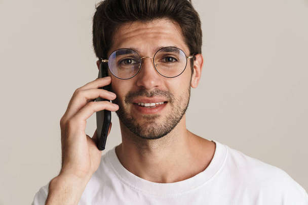 Image of handsome young man in eyeglasses talking on mobile phone isolated by beige background - Valokuva, kuva