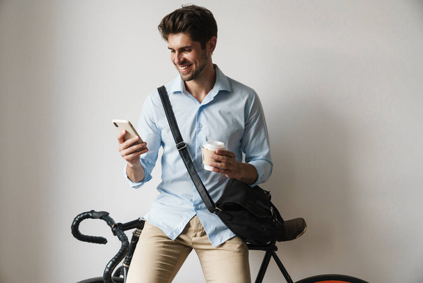 Image of happy businesslike man holding coffee cup and cellphone while standing by bicycle indoors - Foto, immagini