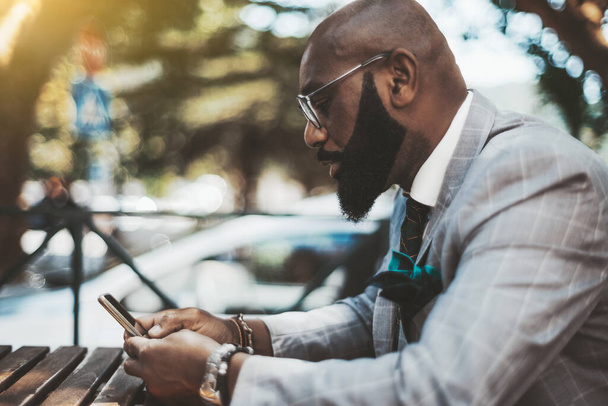 An elegant serious bald mature black guy with a well-groomed beard, in eyeglasses and a fashionable suit, is sitting outdoors at a wooden table of a street bar and using a smartphone to read a message - Фото, зображення
