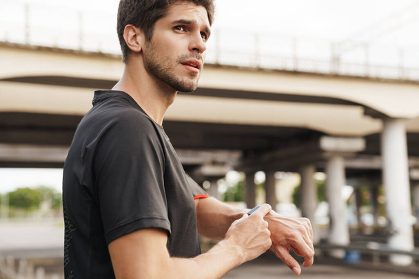 Image of athletic young sportsman using smartwatch while working out on city street - Photo, Image