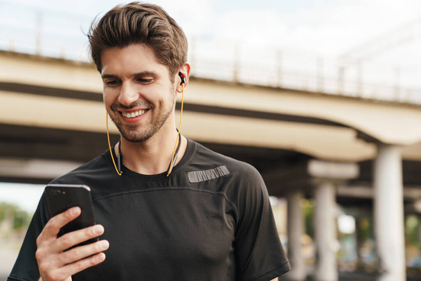 Image of smiling young sportsman in earphones using cellphone while working out on city street - Photo, image