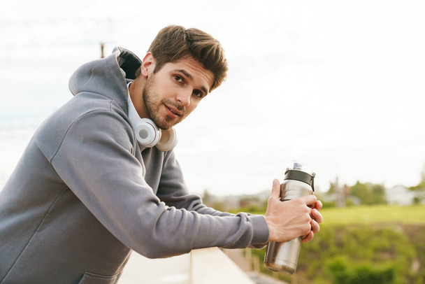 Image of athletic sportsman with headphones holding water bottle while leaning on railing outdoors - Fotó, kép