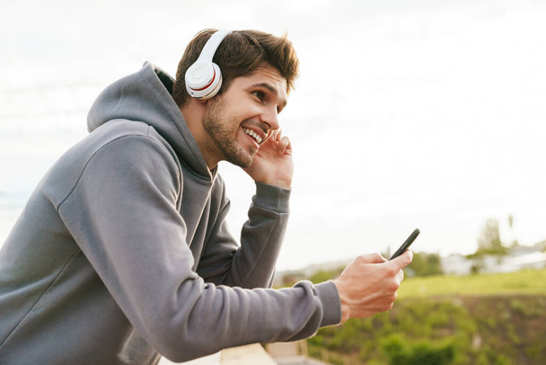 Image of joyful sportsman using headphones and cellphone while leaning on railing outdoors - Foto, afbeelding
