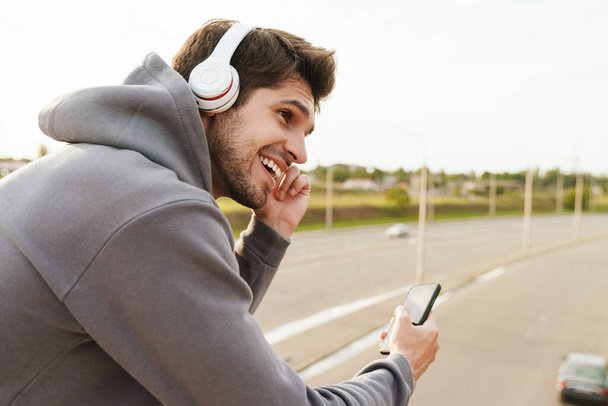 Image of joyful sportsman using headphones and cellphone while leaning on railing outdoors - Photo, image