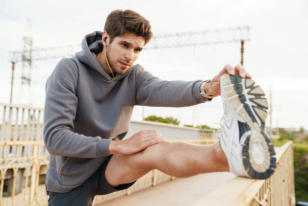 Image of young caucasian sportsman in earphones doing exercise while working out on city bridge - Фото, зображення