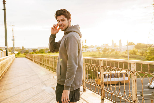 Image of athletic happy sportsman using earphones while working out on city bridge - 写真・画像