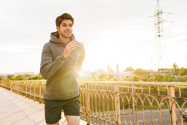 Image of athletic happy sportsman using earphones while running on city bridge - Photo, Image