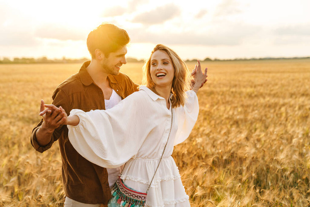 Image of young caucasian beautiful couple holding hands together while walking in golden field on countryside - Photo, Image