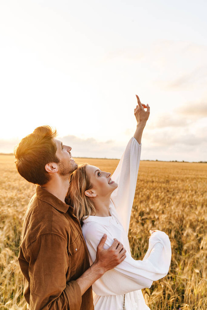 Image of young caucasian couple hugging and gesturing finger upward at sky in golden field - Foto, Bild