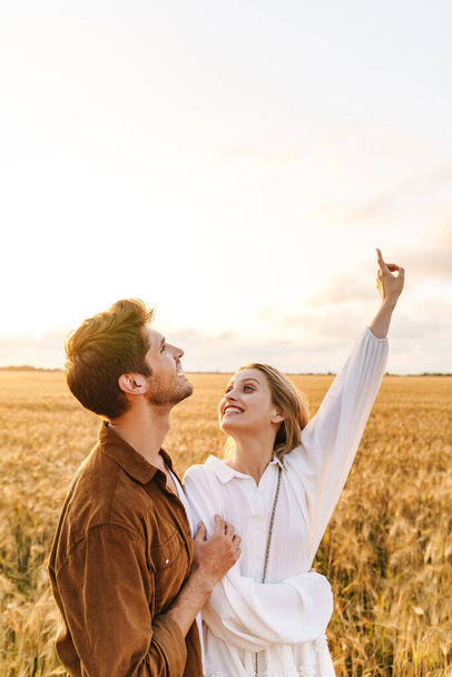 Image of young caucasian couple hugging and gesturing finger upward at sky in golden field - Фото, изображение