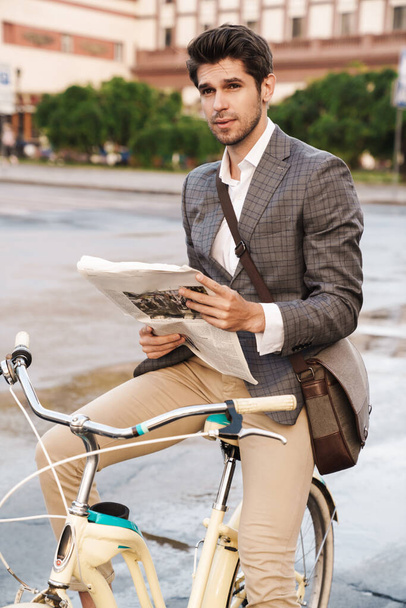 Image of a young serious business man outdoors in park holding newspaper on a bicycle - Photo, Image