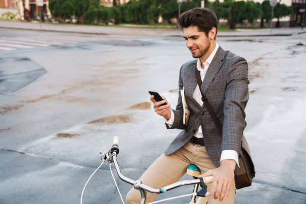 Image of a young handsome pleased smiling business man outdoors using smartphone on a bicycle - Foto, immagini