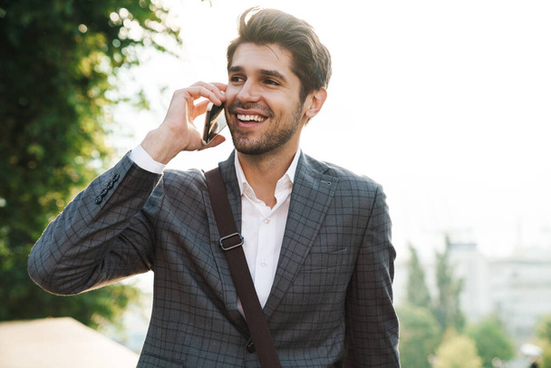 Image of a smiling cheery handsome business man outdoors talking by mobile phone - Zdjęcie, obraz