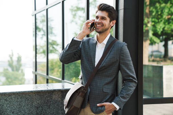 Image of optimistic cheery handsome business man outdoors talking by mobile phone near business center - Photo, Image
