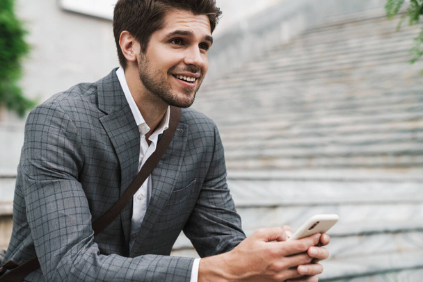 Image of smiling positive handsome business man outdoors using mobile phone while sitting on the stairs - Φωτογραφία, εικόνα
