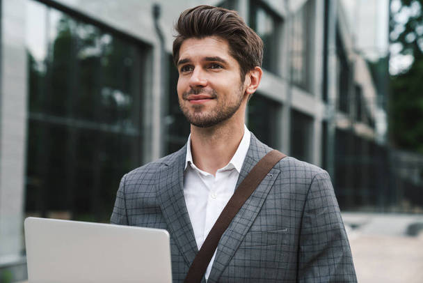 Image of pleased optimistic handsome business man outdoors using laptop computer near business center - Photo, image