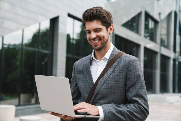 Image of cheerful handsome business man outdoors using laptop computer near business center - Foto, Bild