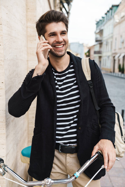 Image of a cheerful young business man outdoors talking by mobile phone on a bicycle - Foto, Imagen