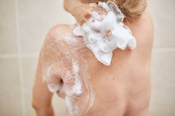 an elderly 60-year-old woman washes in the shower, Soaps her shoulders, back and body with a washcloth in the home bathroom. selective focus, small focus area. - Foto, Imagen