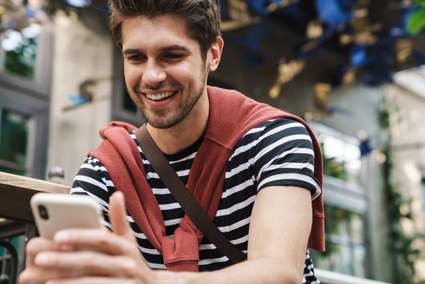 Image of cheerful handsome man smiling and using cellphone while walking on city street - Fotoğraf, Görsel