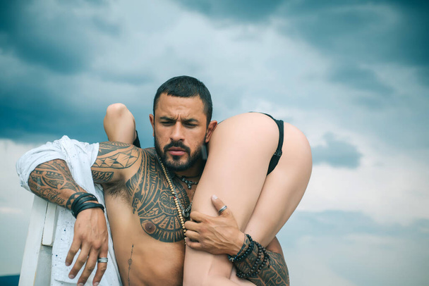 Man holding woman with sexy hot ass. Strong muscular tattooed bearded man holding his sexy fit woman with hot butt isolated at grey background. Eternal love and trust concept. - Zdjęcie, obraz