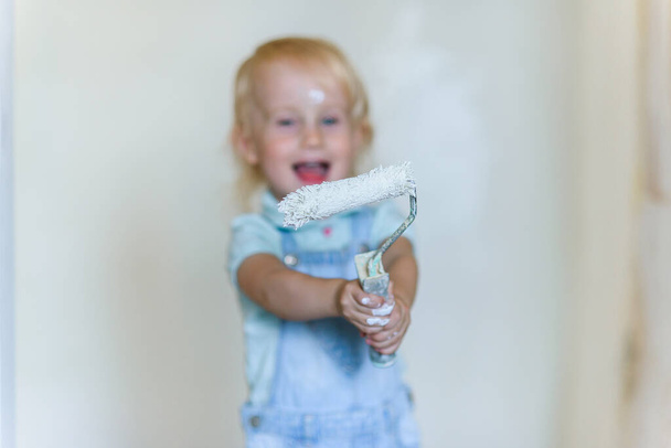Funny toddler girl holds a white paint roller and smiling in a new house. Selective focus. - Foto, immagini