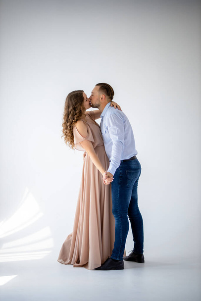 A beautiful curly pregnant girl in an evening light  dress kissing her husband on a white background. She has a long evening dress and he is wearing festive blue shirt. Family and Happiness Concept - Foto, afbeelding