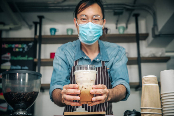 Zakenman draagt gezichtsmasker voor goede hygiëne koffiehuis eigenaar serveert ijs koffie jonge ondernemer, vriendelijke serveerster met een bril glimlachend vasthouden en serveren. - Foto, afbeelding