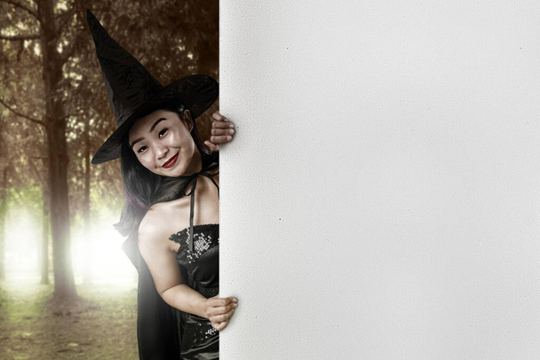 Asian witch woman with hat standing with empty whiteboard with haunted forest background. Empty whiteboard for copy space - Foto, immagini