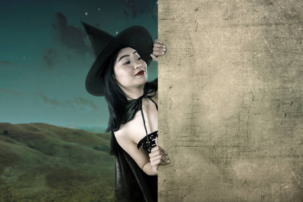 Asian witch woman with a hat standing with an empty board with a night scene background. Empty board for copy space - Foto, Imagen