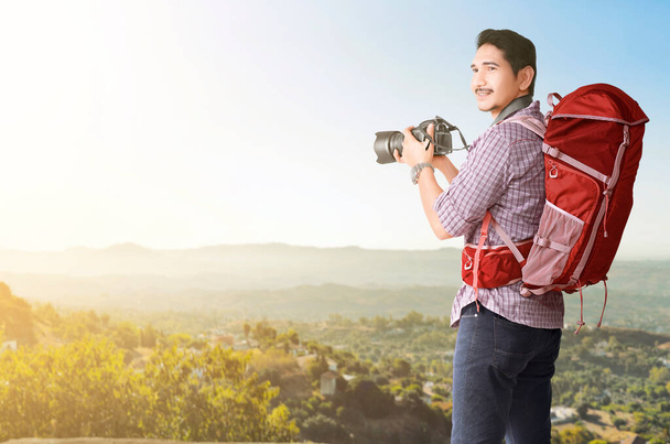 Asian man with a backpack holding camera with a mountain view and blue sky background. World Tourism Day. - Fotografie, Obrázek