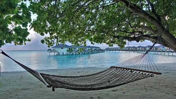 Manhã nas Maldivas. Há uma rede na praia entre as árvores. Ramos dobrados sobre a areia. Uma fileira de moradias aquáticas pode ser vista acima do oceano aquamarino. Nuvens no céu. - Foto, Imagem