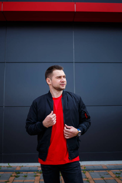 A young man dressed in urban style stands on the street against a gray wall, he is in a black jacket and a red T-shirt. Stylish guy's orchestra is the background for your promotional text message. - Foto, Imagen