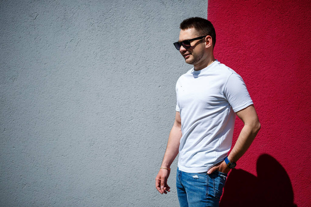 Retrato de um jovem elegante, um homem vestido com uma camiseta branca em branco em pé sobre um fundo de parede cinza e vermelho. Estilo urbano de roupa, imagem na moda moderna. Moda masculina - Foto, Imagem
