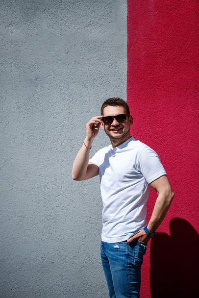 Портрет стильного молодого чоловіка, чоловіка, одягненого в білу порожню футболку, що стоїть на сірому і червоному тлі стіни. Міський стиль одягу, сучасний модний образ. Чоловіча мода
 - Фото, зображення