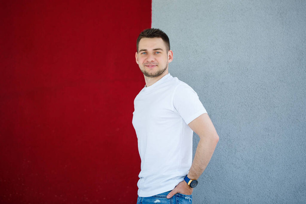 Stylish young man, a man dressed in a white blank t-shirt standing on a gray and red wall background. Urban style of clothes, modern fashionable image. Men's fashion - Photo, Image
