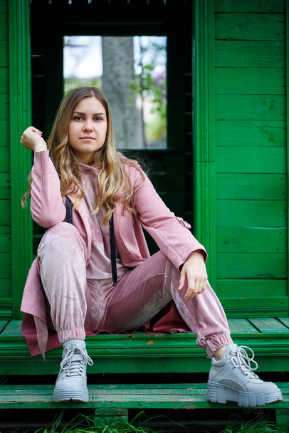 Stylish young girl with blond hair of European appearance sits on the steps, dressed in a pink suit, modern womens fashion, casual clothes - Foto, Imagen