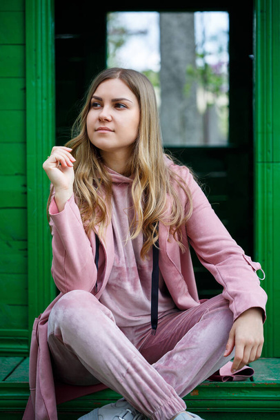 Stylish young girl with blond hair of European appearance sits on the steps, dressed in a pink suit, modern womens fashion, casual clothes - Φωτογραφία, εικόνα