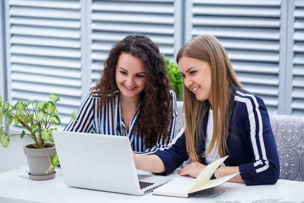 Duas jovens empresárias bem sucedidas estão sentadas em uma mesa com um laptop e um notebook trabalhando em um novo projeto de desenvolvimento, estudantes do sexo feminino escrever um relatório sobre o trabalho de seu computador - Foto, Imagem