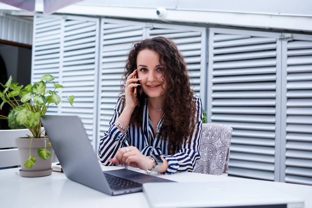 Улыбающаяся успешная молодая женщина разговаривает по телефону, работает на ноутбуке на террасе, девушка в кафе на деловой встрече. Концепция бизнеса - Фото, изображение