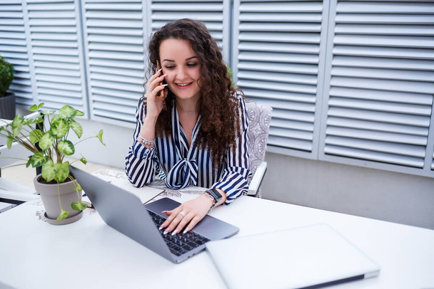 Hymyilevä nuori nainen puhuu puhelimessa, työskentelee kannettavan tietokoneen terassilla, tyttö kahvilassa liiketapaamisessa. Liiketoiminta - Valokuva, kuva
