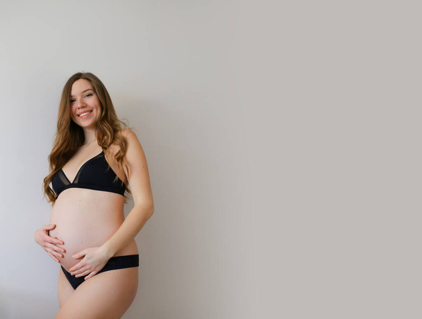 Siyah iç çamaşırlı hamile bir Amerikalı kadın. Gri fotokopi arka planında karnını tutuyor.. - Fotoğraf, Görsel